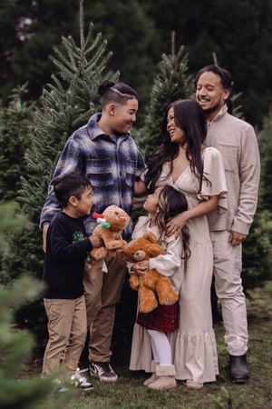 Family poses together at Christmas tree farm with teddy bear in coordinating neutral outfits.