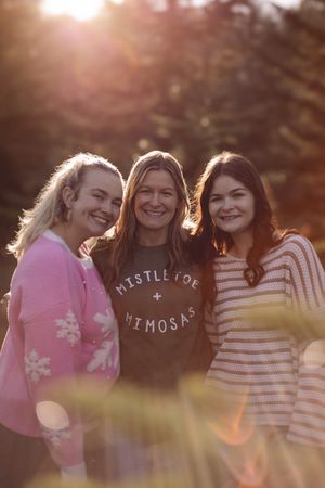 Three friends smile together outdoors at sunset wearing cozy sweaters in pink, black and striped patterns.