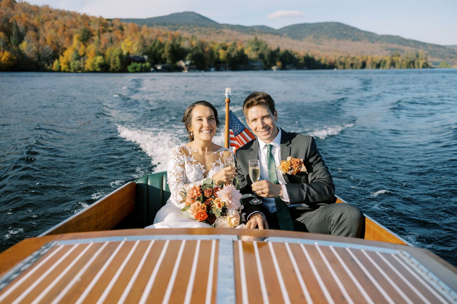 A Chris-Craft boat glides across Lake Placid during autumn with fall foliage reflecting in the calm waters and the bride and groom cheersing