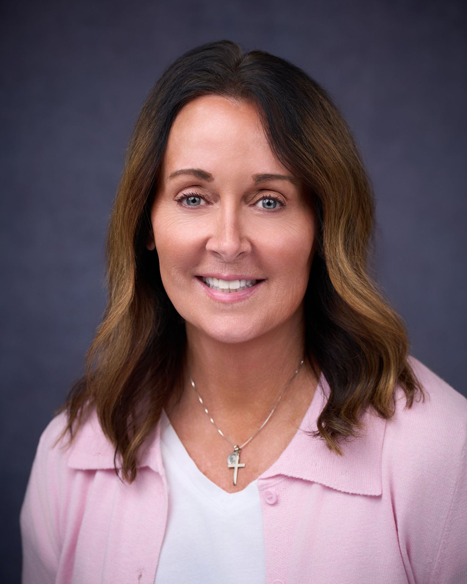 Professional headshot of a smiling woman with brown hair wearing a pink blazer and cross necklace.