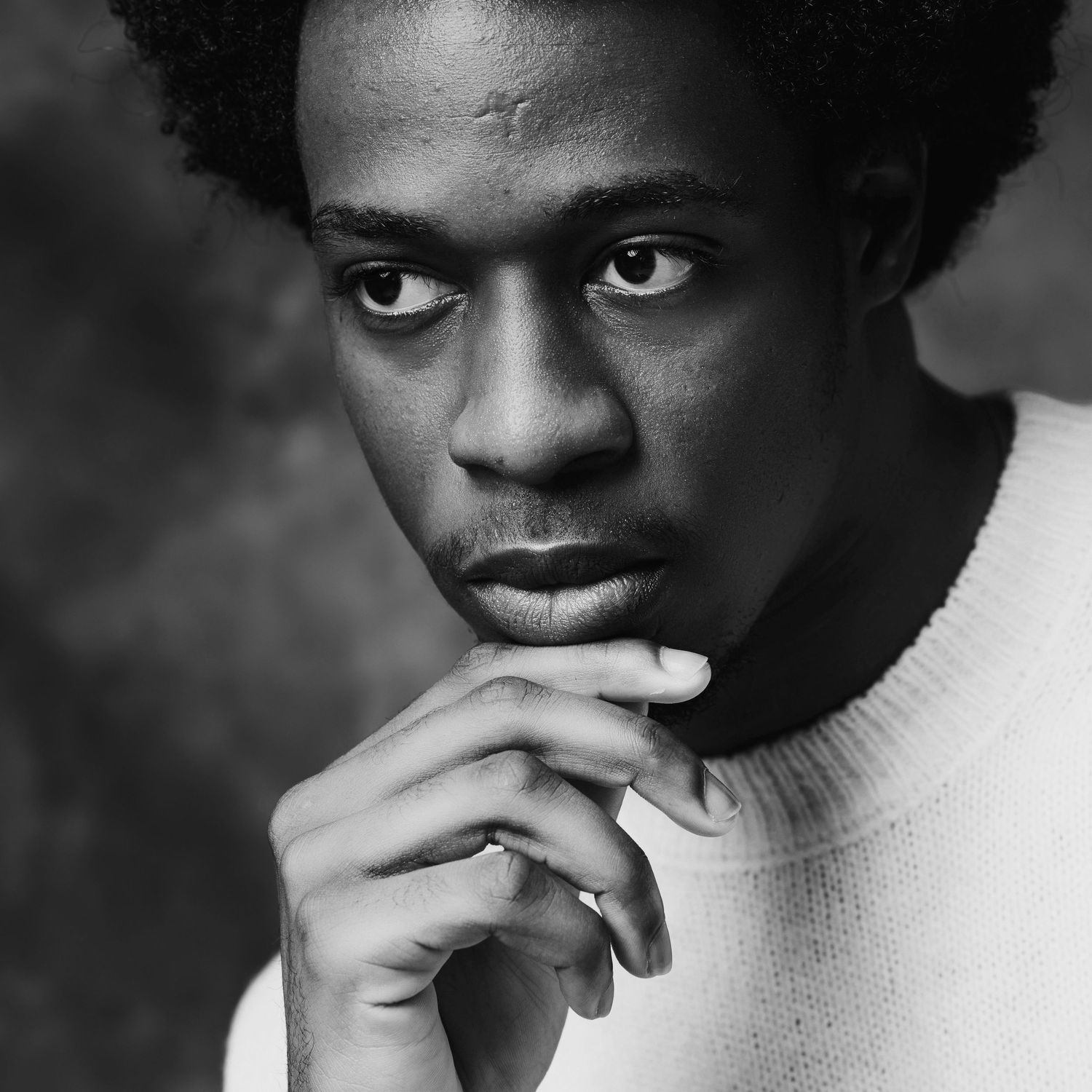 Black and white headshot portrait of a young man in a white sweater, chin resting on hands, with a thoughtful expression.