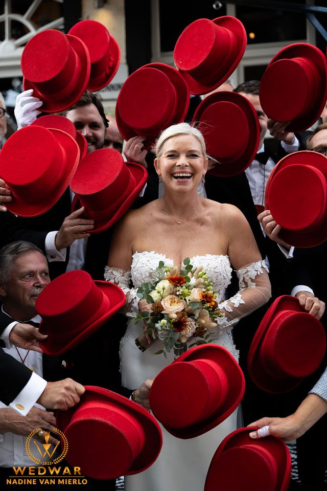 A bride in a white strapless dress stands surrounded by guests holding bright red top hats at a wedding celebration.