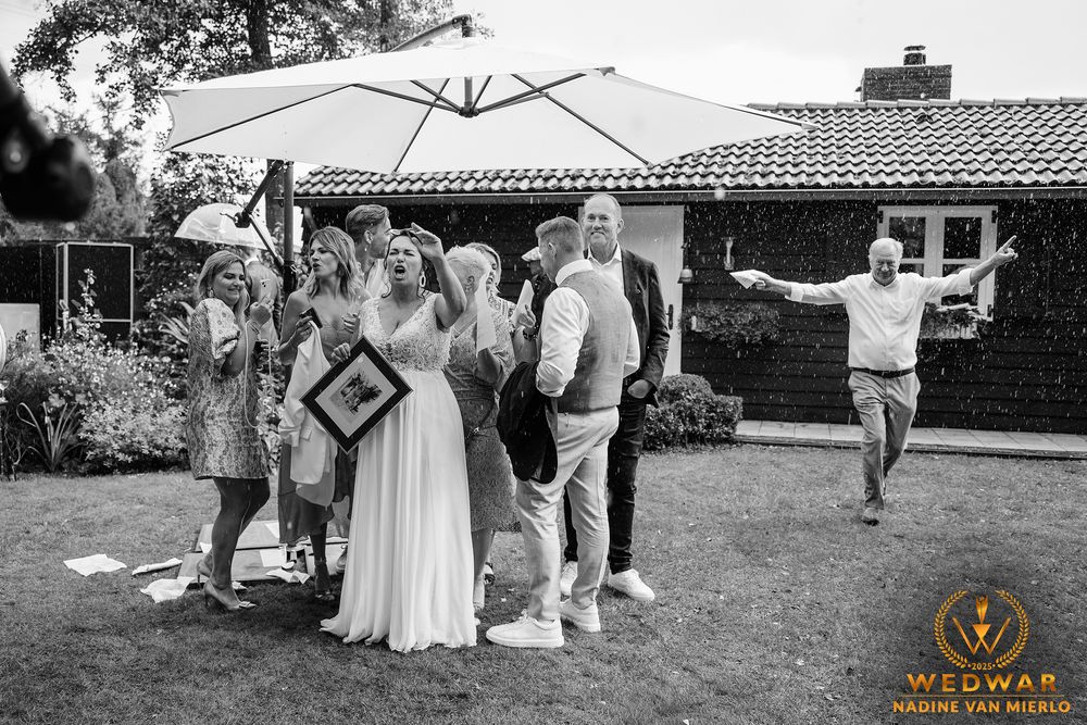 Black and white photo of outdoor wedding guests gathered under a patio umbrella while someone dances in the background.