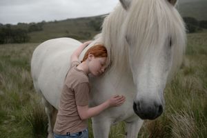 A person in a peach colored shirt tenderly embraces a white horse in a grassy meadow setting.