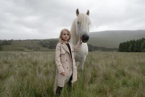 A beautiful white horse stands in a misty field beside someone wearing a beige coat on a cloudy day.