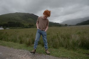 A person walking through misty Scottish highlands with dramatic cloudy skies and green rolling hills in the background.