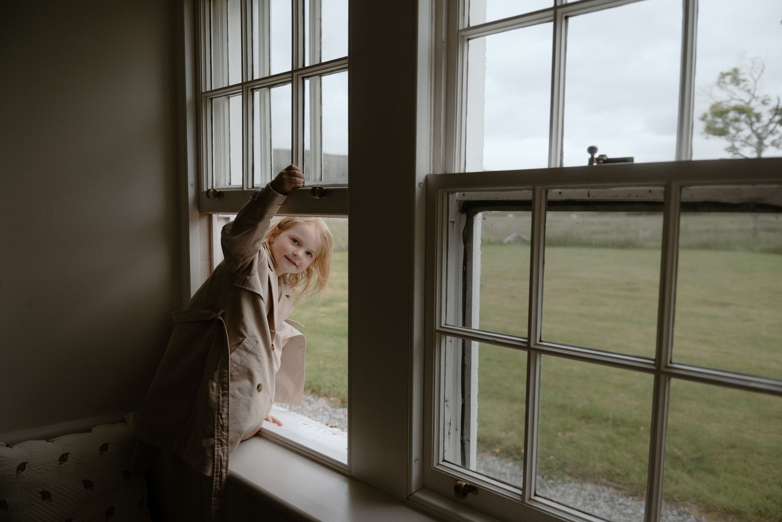 A person in a white dress gazes through a window overlooking a serene countryside landscape.