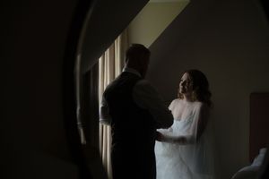 A couple stands together in silhouette by a window in a dimly lit room during an intimate wedding moment.