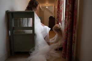 A bride in a flowing white wedding gown walks down a hallway with natural light streaming through windows.