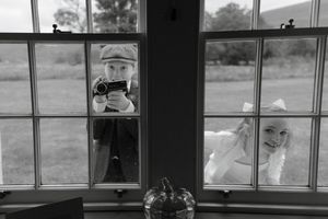 Black and white photo showing someone taking a picture through a window while another figure peeks from an adjacent pane.
