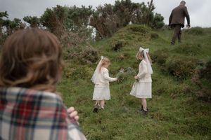 People in light colored dresses walk up a grassy hillside while someone in plaid watches from a distance.