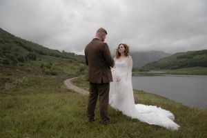 A couple stands together on a misty hillside overlooking a scenic Scottish loch on their wedding day.