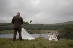Wedding party relaxes on a grassy hillside overlooking a serene lake on an overcast day in the countryside.