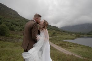 A couple shares an intimate moment in the scenic Scottish Highlands with misty mountains and a loch in the background.