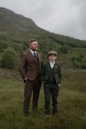 Two figures stand in rugged Scottish countryside wearing brown and dark green suits against misty hills.