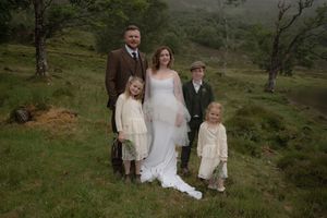 Family members stand together in a rustic outdoor setting with rolling green hills and trees in the background.