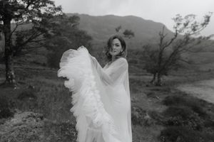 A figure in a flowing white dress and veil stands in a dramatic black and white landscape with mountains in the background.