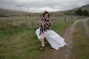 A person in a white dress walks along a winding dirt path through rolling green hills under cloudy skies.