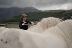 A person rides a white horse along a misty mountain trail with dramatic cloudy skies in the background.