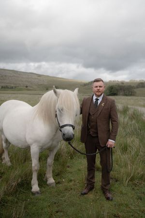 White Highland pony grazes in misty Scottish moorland alongside someone in a brown tweed suit.