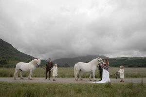 A romantic wedding party poses with two majestic white horses against a misty Scottish Highland landscape.