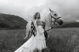 A bride in a flowing white dress poses with a majestic horse in a scenic mountain meadow during a photoshoot.