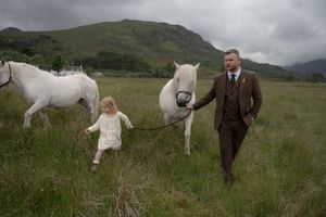 A white horse runs playfully through a green field while being watched by people in traditional country attire.
