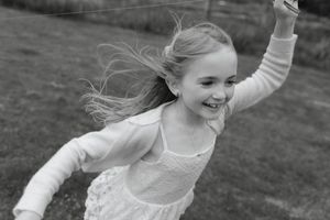A young dancer in a white dress twirls joyfully outdoors with windswept hair in black and white.