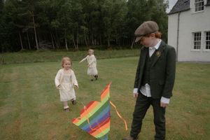 Children in vintage-style clothing play with a colorful rainbow flag on a grassy lawn near a white house.