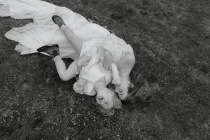 A child in white clothing lies on dark grass while looking upward at the camera.