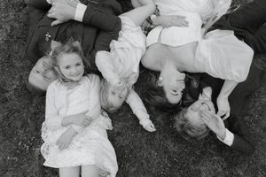 Multiple children in white clothing lie on dark grass while looking upward at the camera.
