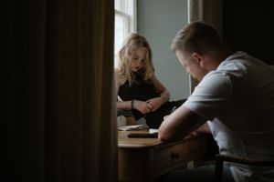 Students study together at wooden tables near windows in a dimly lit library setting.