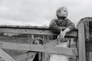 A child hangs playfully on a wooden fence rail in a black and white countryside setting.