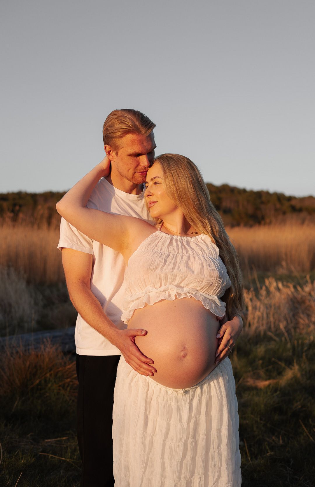 gravid kvinna står lutad emot en man, han håller om hennes mage. kvinnan kollar bortåt och mannen på henne.