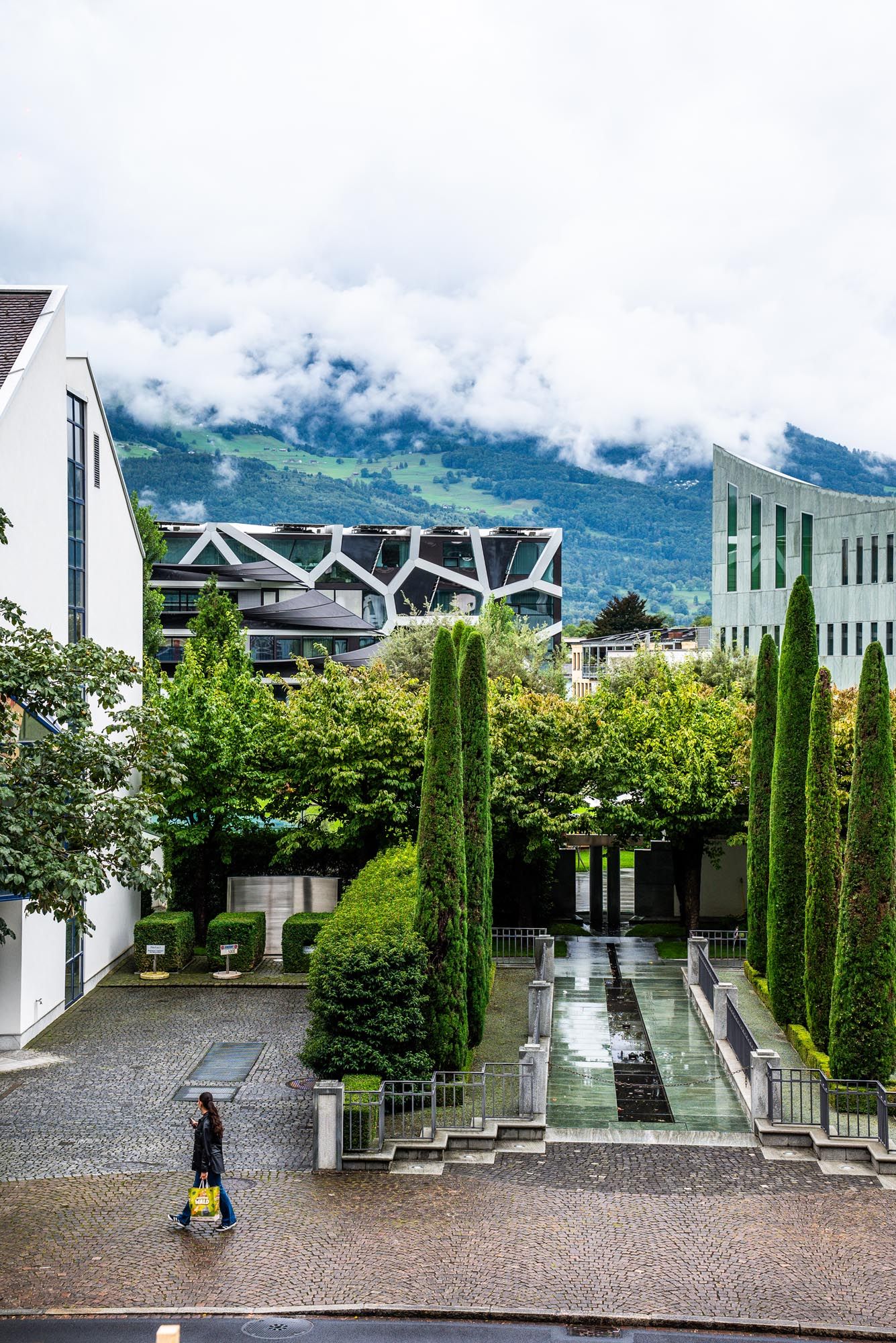 Modern courtyard plaza with tall cypress trees and white columns against misty mountain backdrop in European setting.