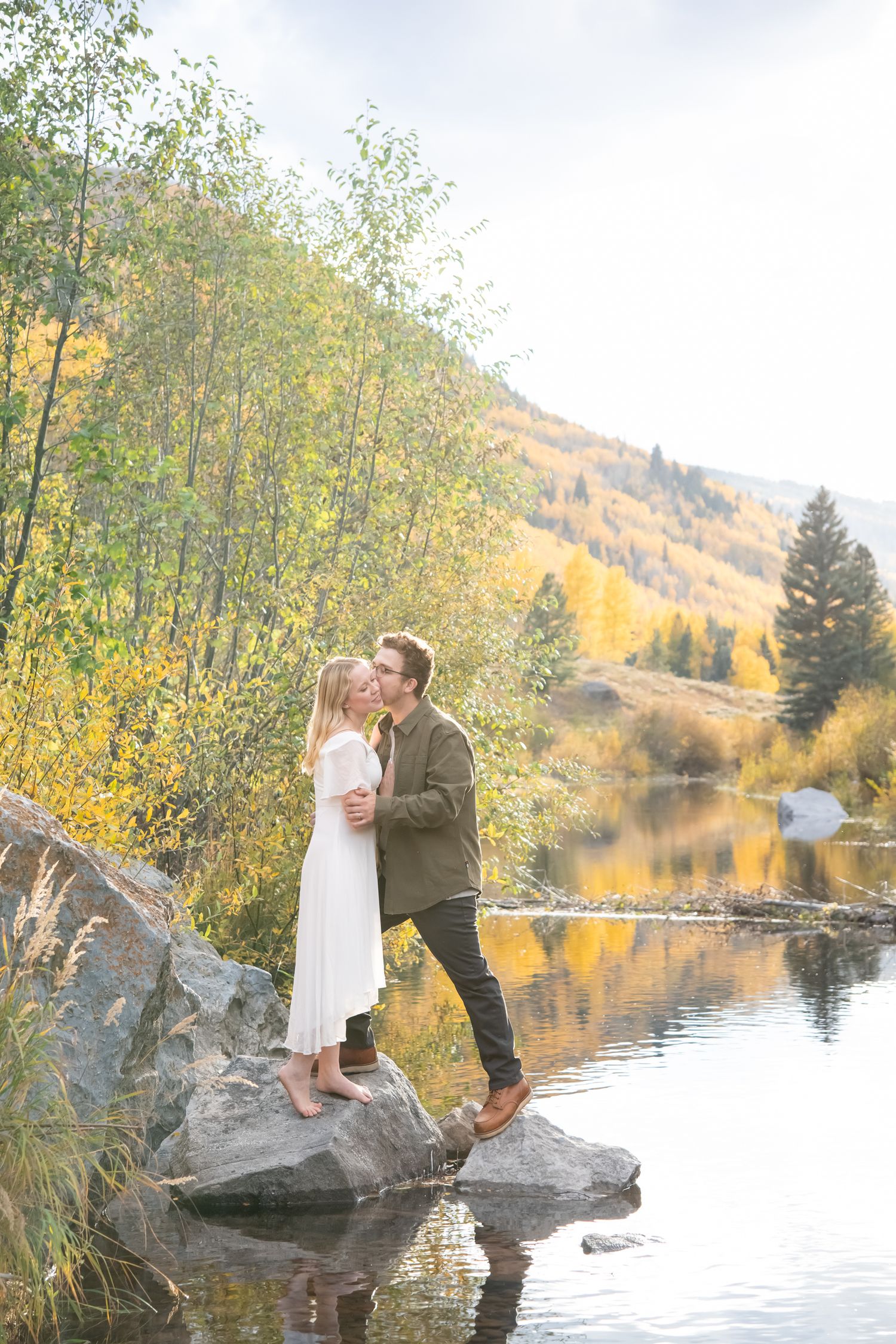 Beautiful couple kisses while standing on a rock in the middle of the creek with fall foliage and aspen trees on the mountains behind them