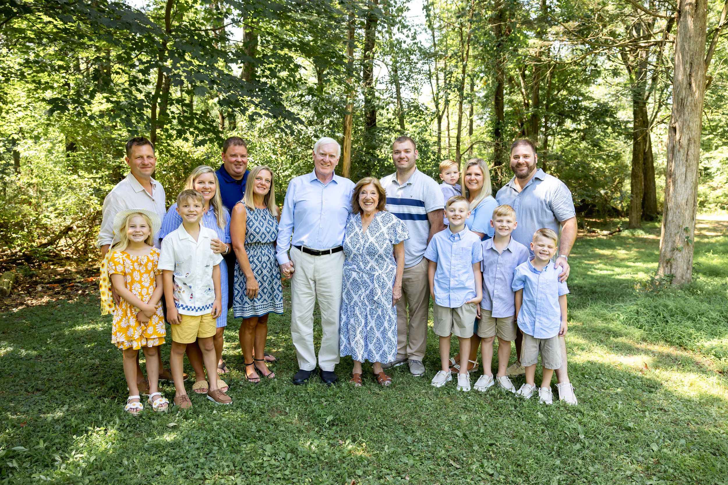 Large family group wearing coordinated blue and yellow outfits poses together in a sunny wooded outdoor setting.