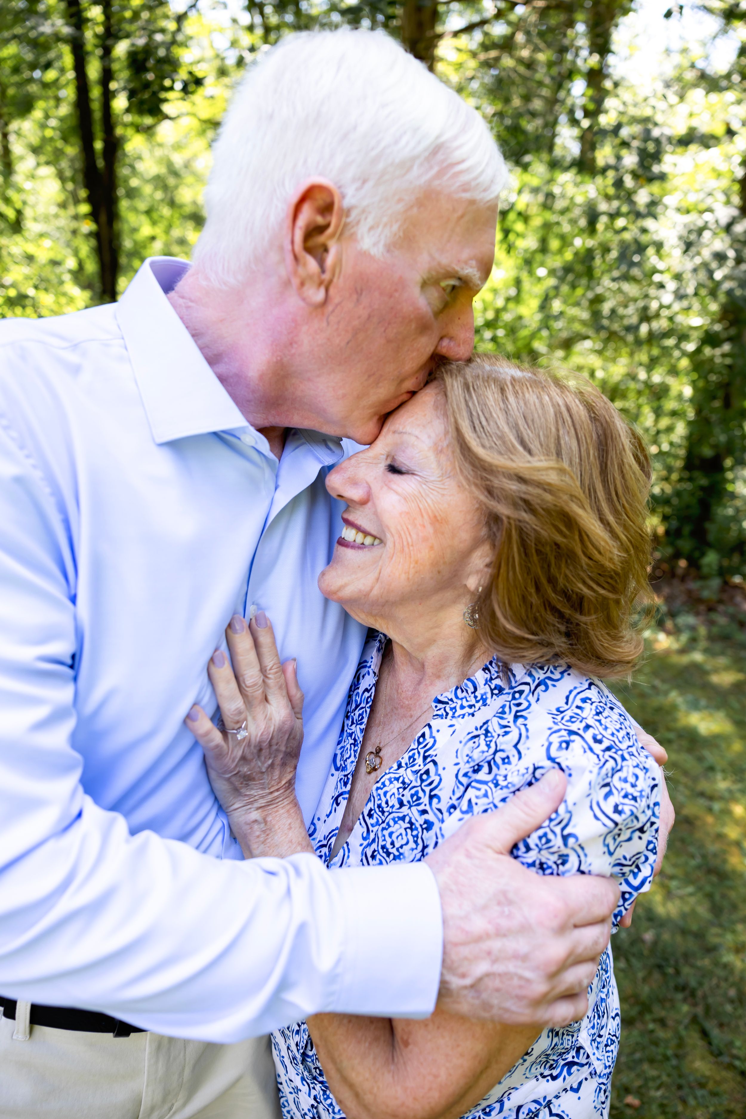Couple in blue and white outfits share intimate moments in a sunlit forest setting.