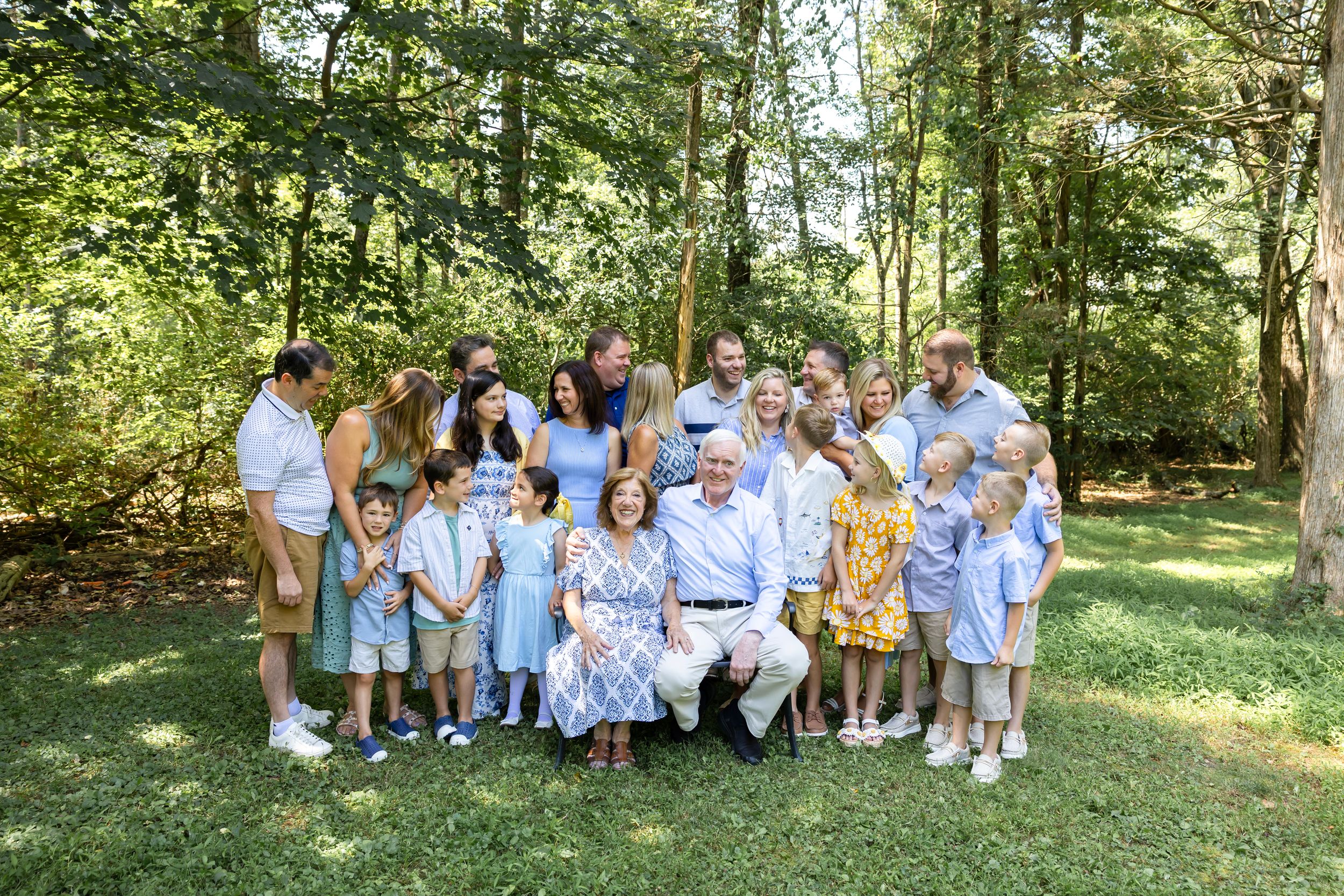Extended family group photo session in a sunlit wooded area wearing coordinated blue and white attire.