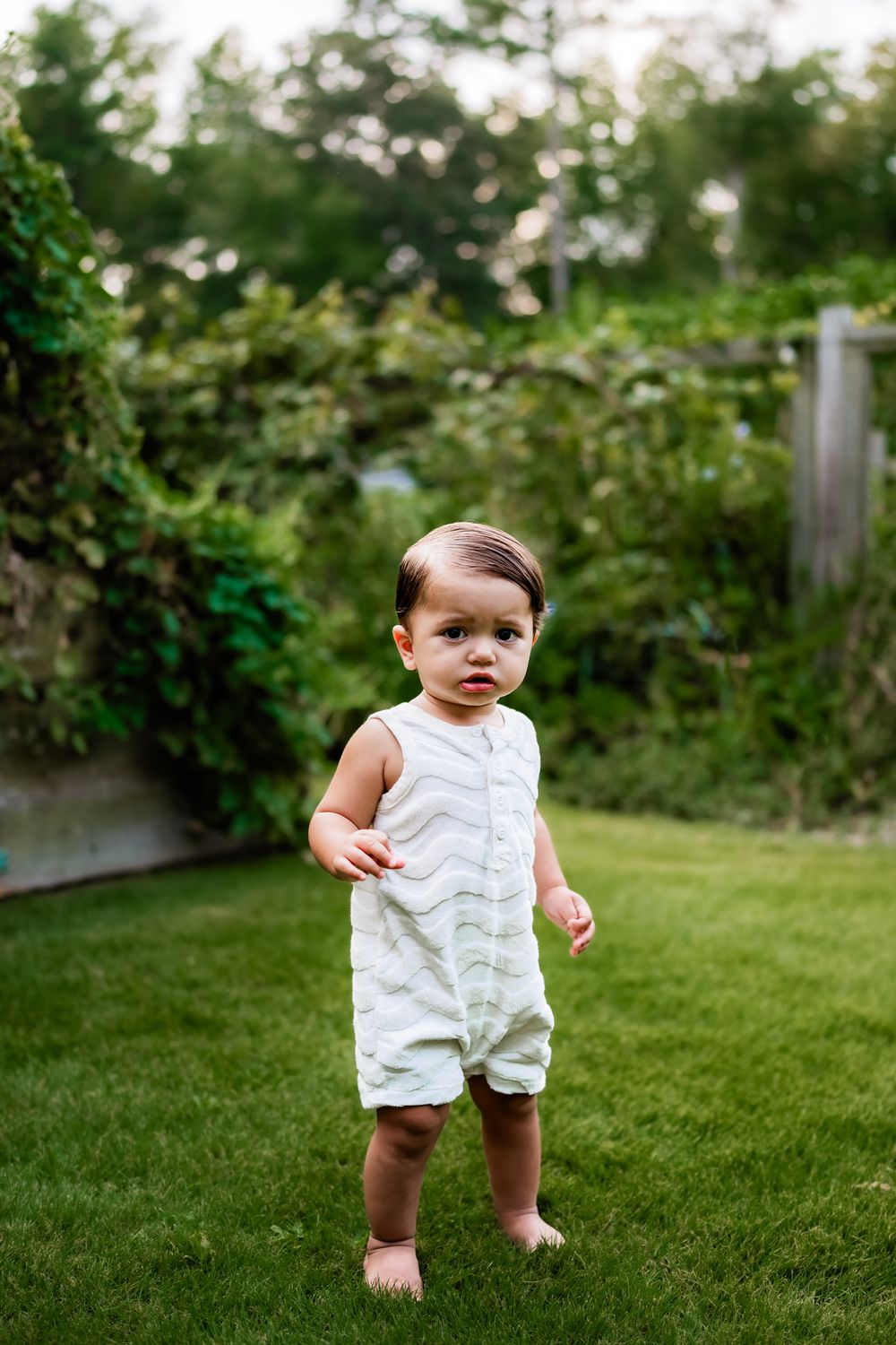 A small child wearing a white sleeveless romper stands barefoot on green grass in a garden setting.
