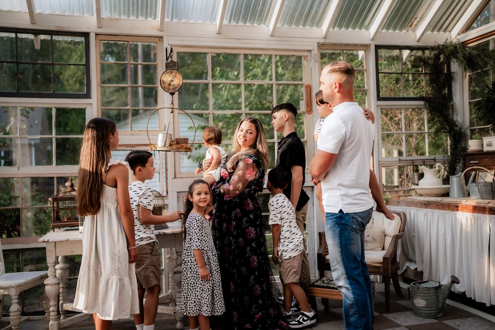 A group of wedding guests gather in a bright greenhouse venue with glass walls and natural lighting.