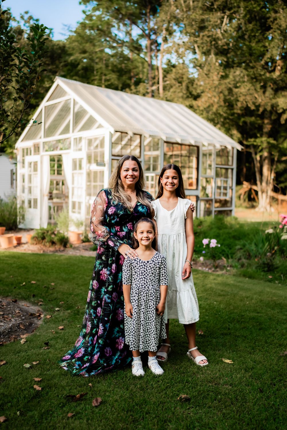 Three people stand together smiling in front of a white greenhouse structure in a garden setting.