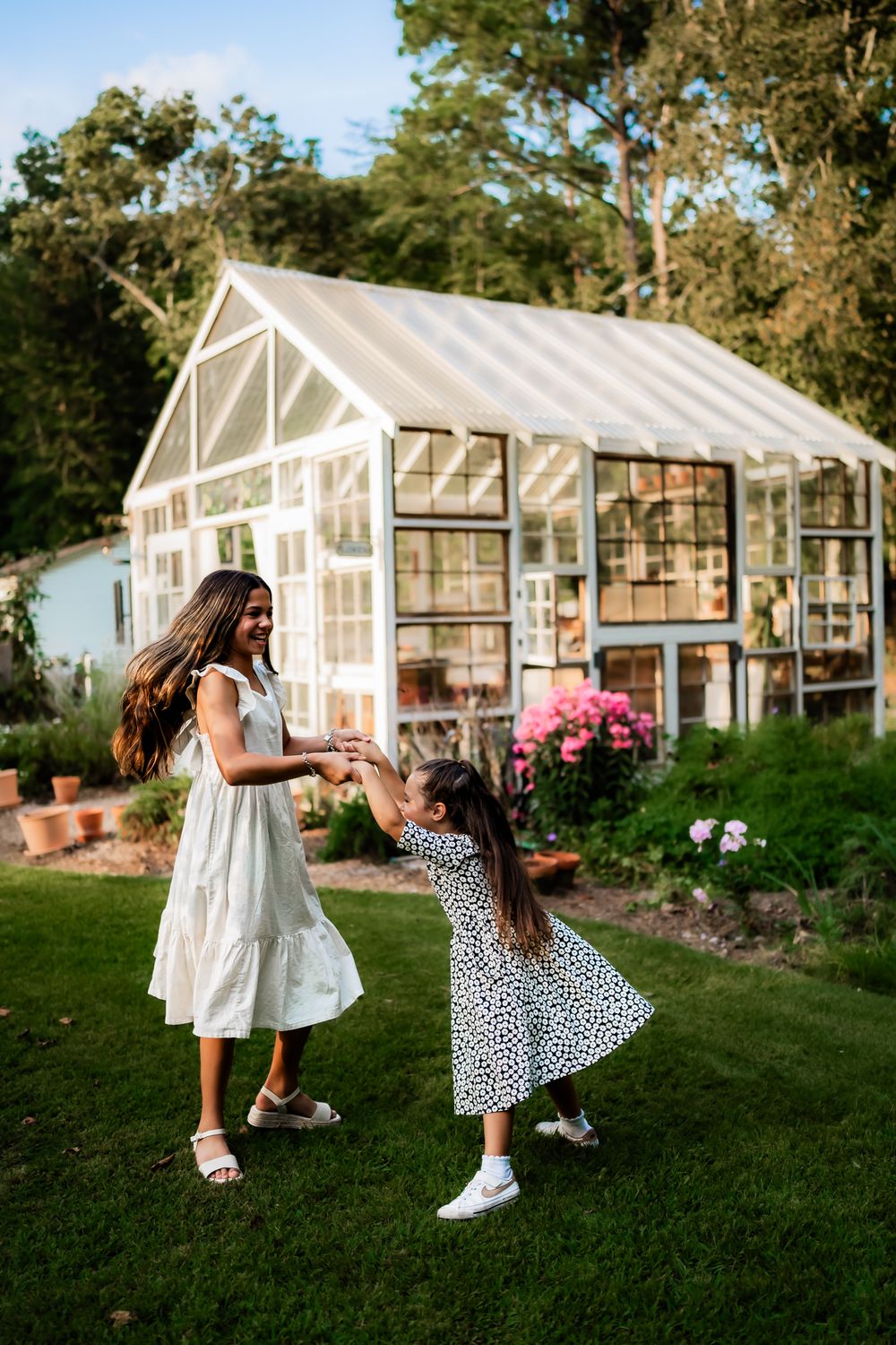 Two people dance and play together on a green lawn in front of white greenhouse structures on a sunny afternoon.
