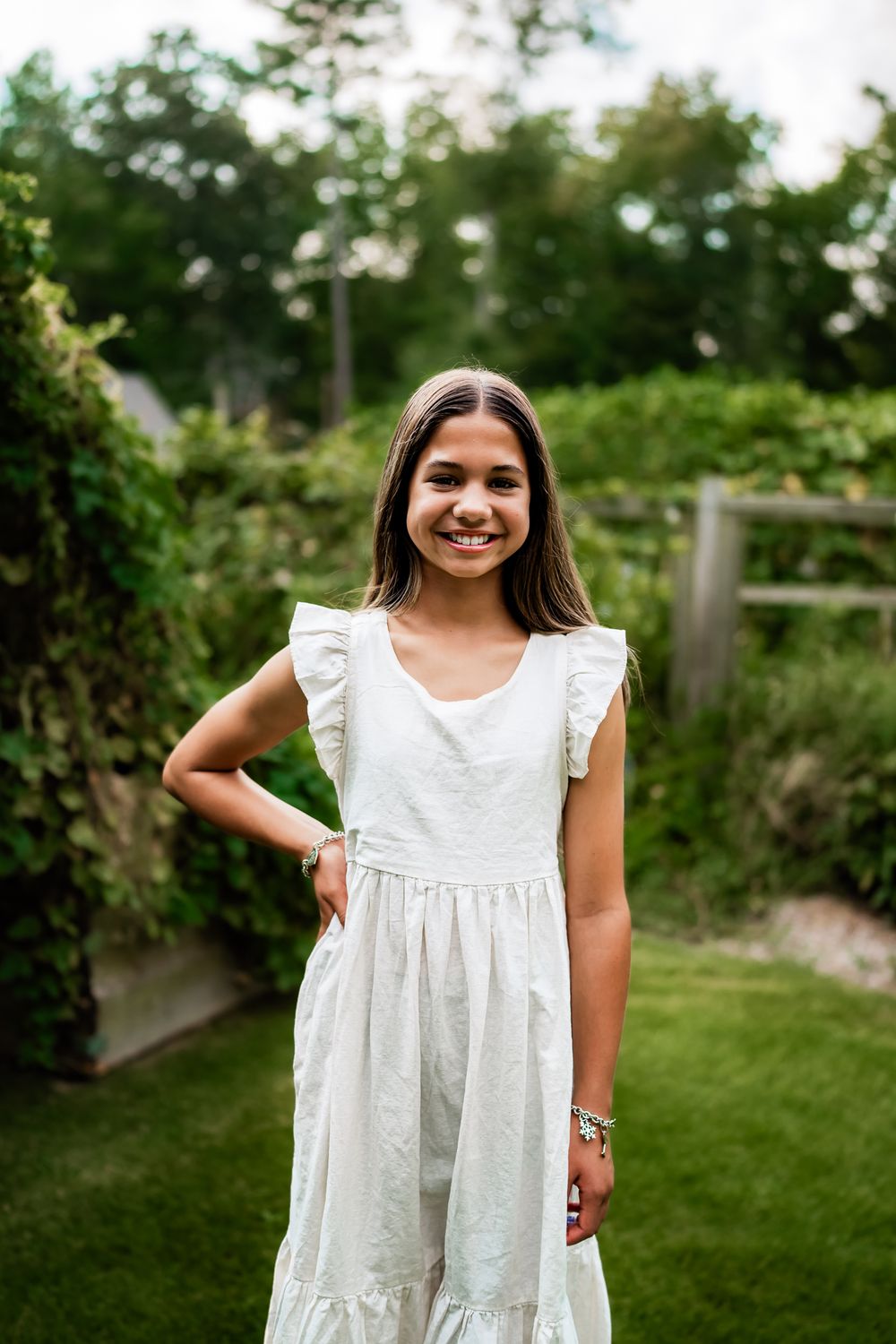 Young person in a white summer dress standing in a sunny backyard garden setting.