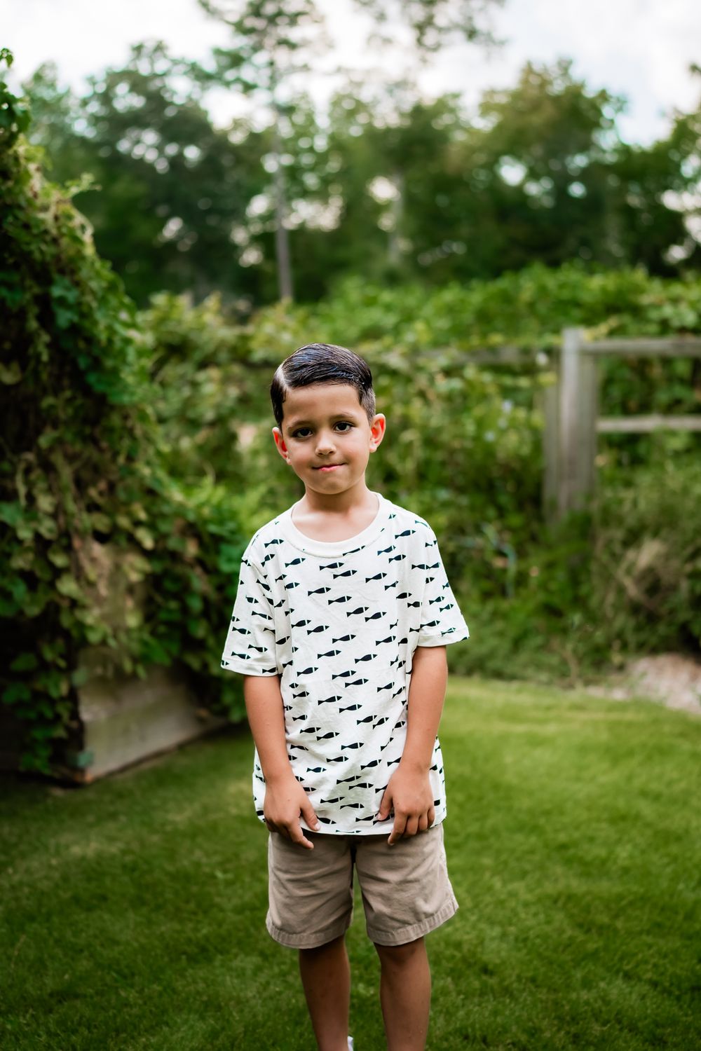 A child stands in a backyard garden wearing a patterned t-shirt and khaki shorts on a sunny summer day.