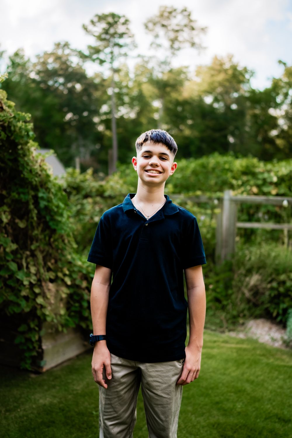Portrait of a person in a black polo shirt and gray pants standing in a backyard garden setting at dusk.
