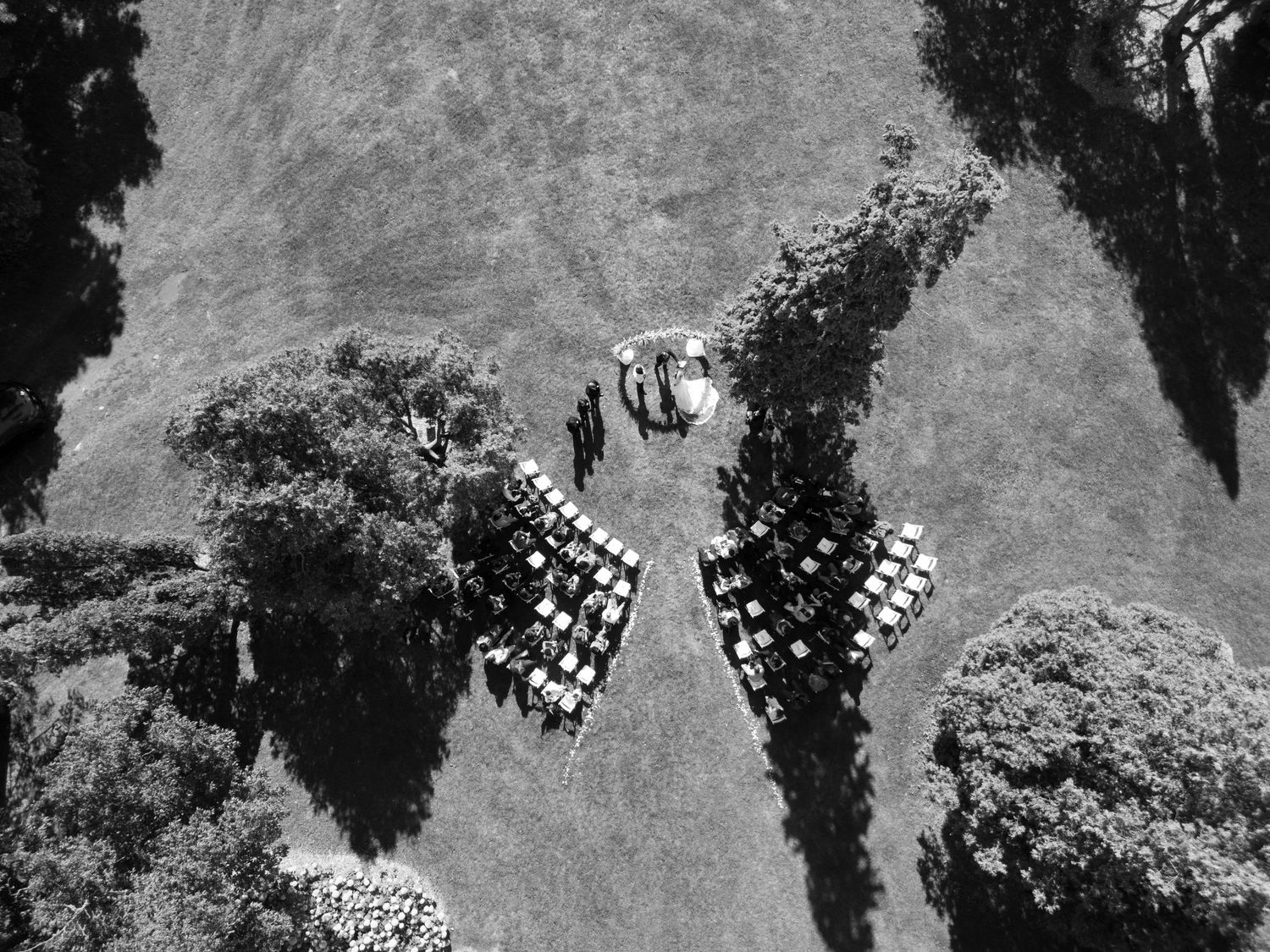 Aerial black and white view of an outdoor wedding ceremony with rows of chairs arranged on a grassy hillside.