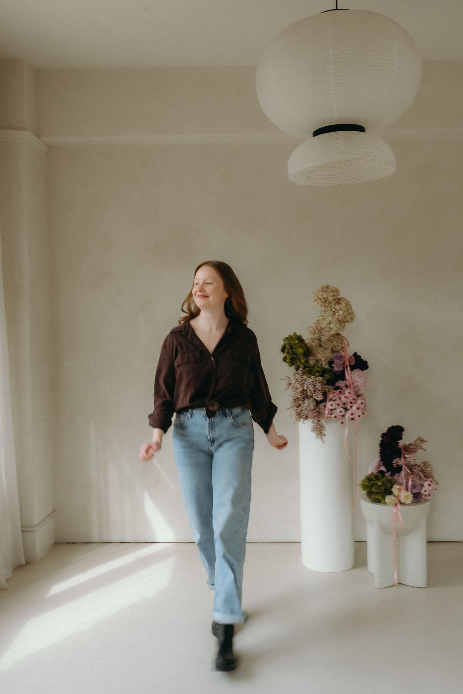 A person wearing jeans and dark top dances across a minimalist white room with soft lighting and floral arrangements.