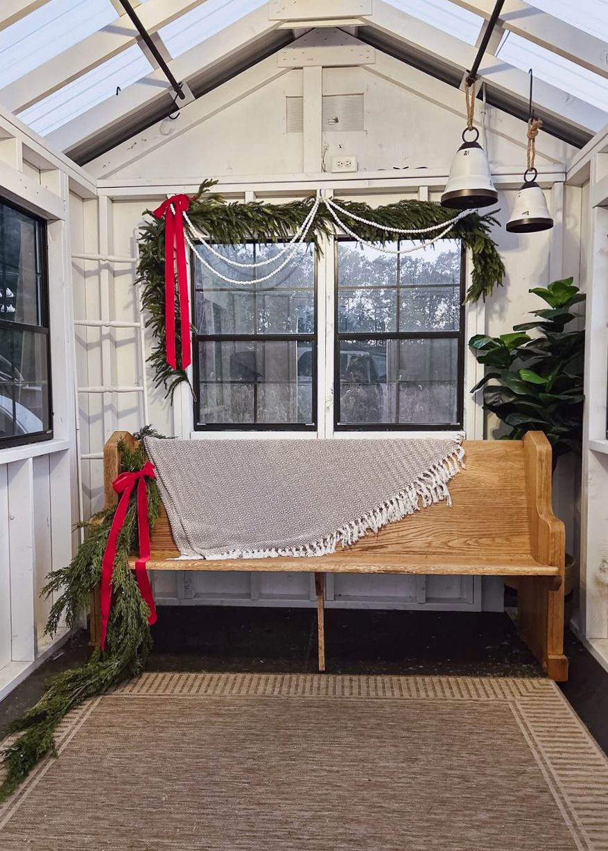 A cozy greenhouse sunroom with a wooden bench, macrame hammock, and festive red ribbons and greenery decorations.