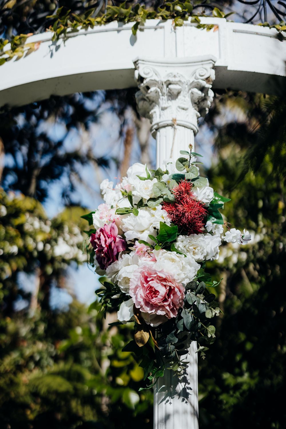White and pink floral arrangements decorate ornate white pillars in an outdoor garden setting.